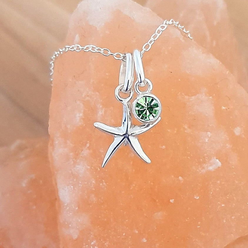 A sterling silver starfish pendant necklace with a green birthstone crystal, displayed on a orange textured surface.