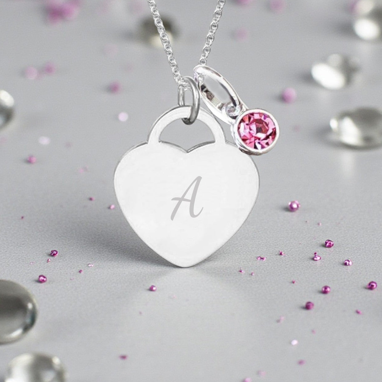 Silver necklace with a heart-shaped pendant and pink gemstone on a gray background with scattered gemstones.