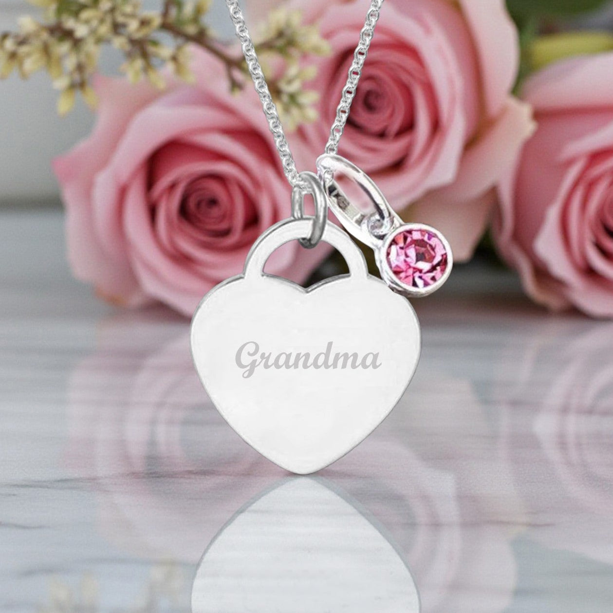 Heart-shaped necklace with 'Grandma' engraving and pink gemstone in front of pink roses.