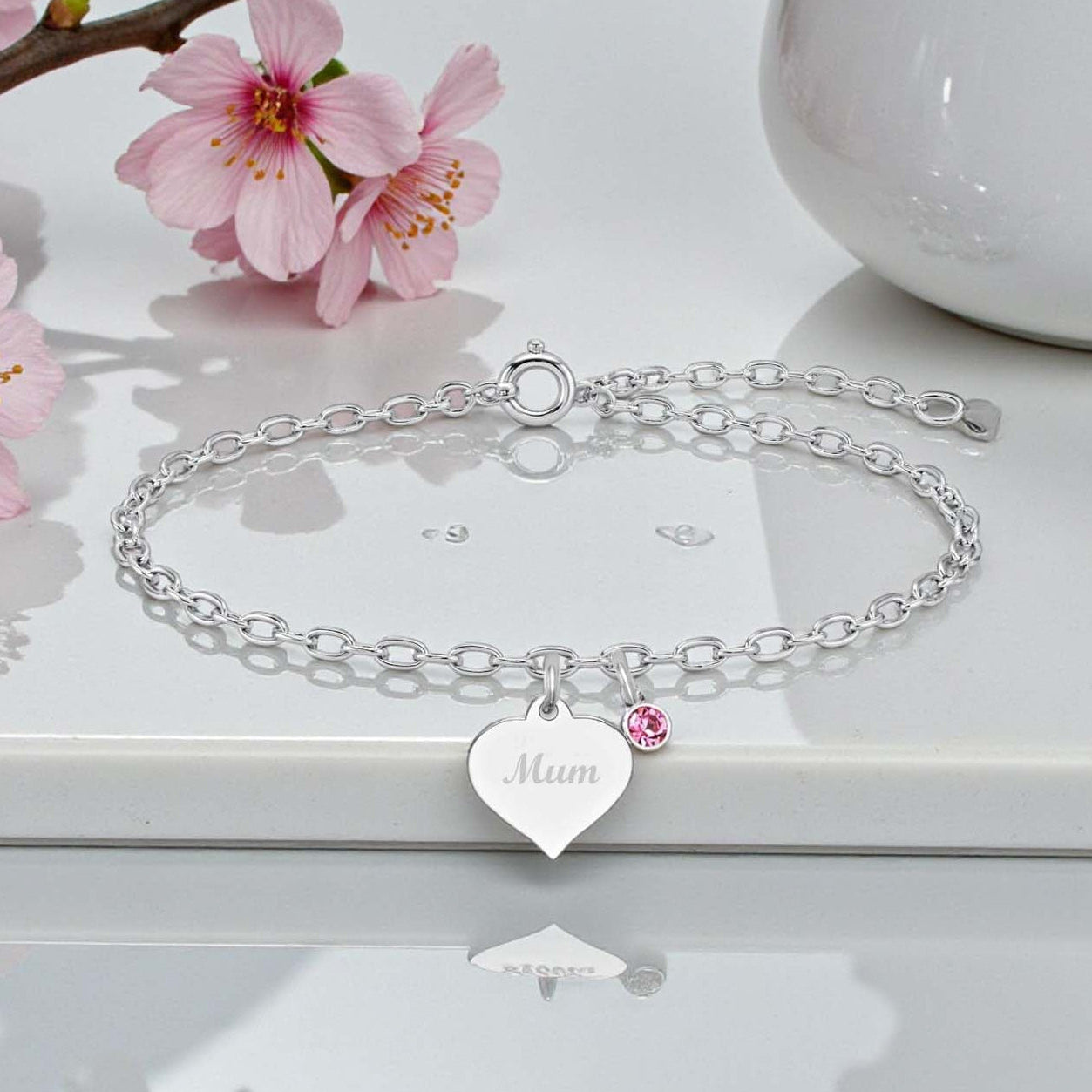 Silver bracelet with a heart-shaped 'Mum' tag on a white surface with cherry blossoms.