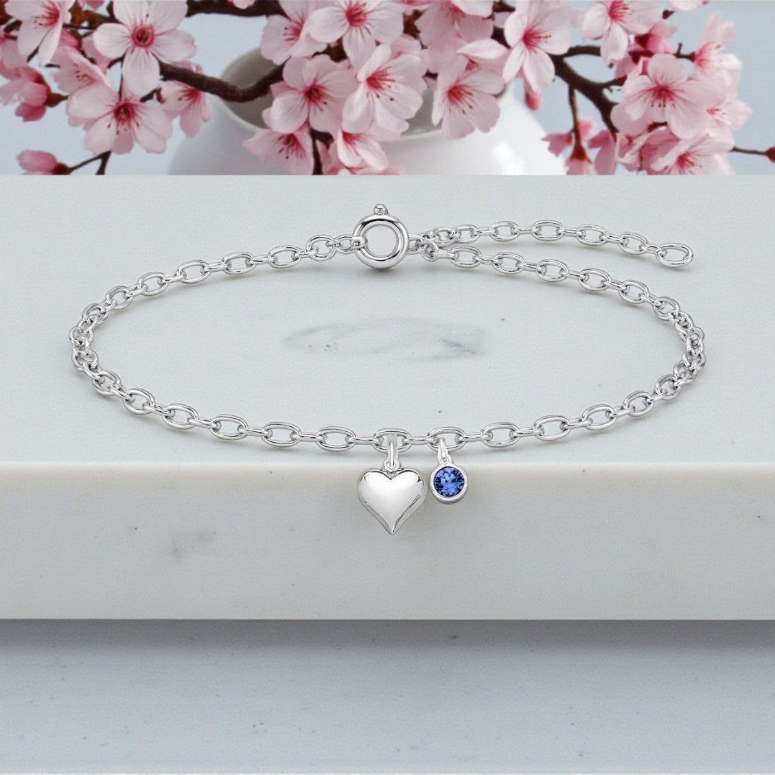 Silver bracelet with heart and blue gemstone charm on a marble surface with cherry blossoms in the background